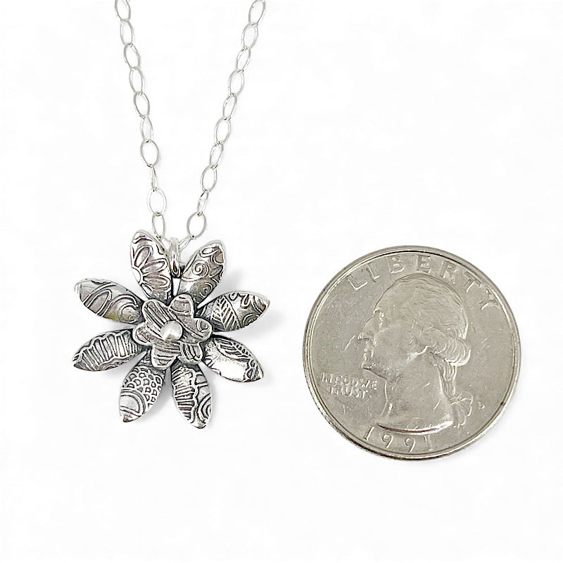 Silver flower-shaped pendant on a chain next to a quarter for scale on a white background